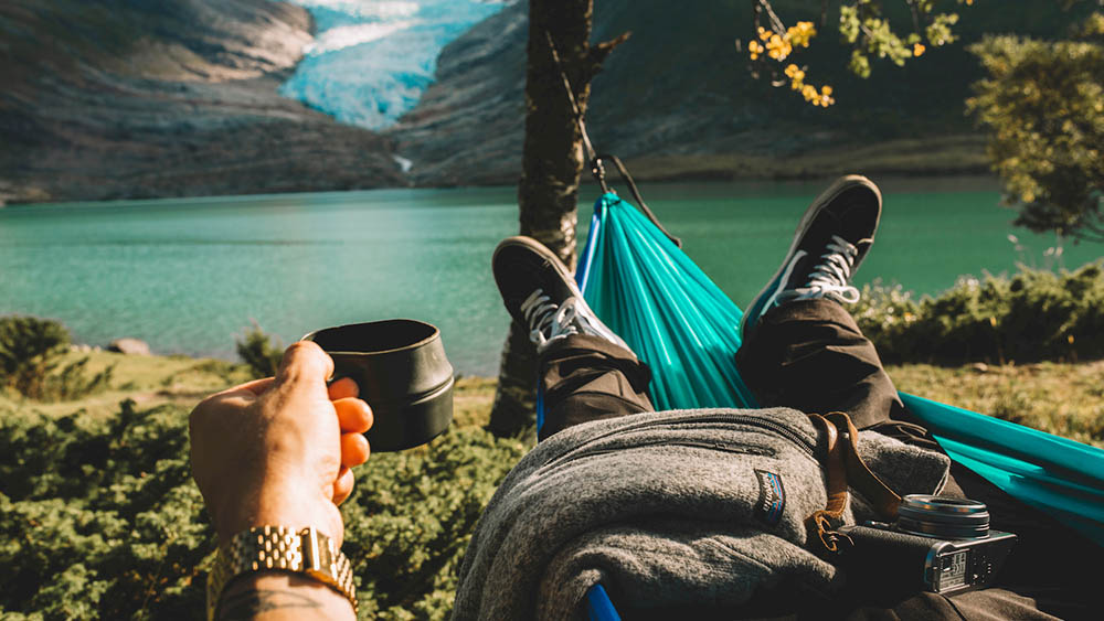 Man with coffee in hammock next to lake and glacier in Norway