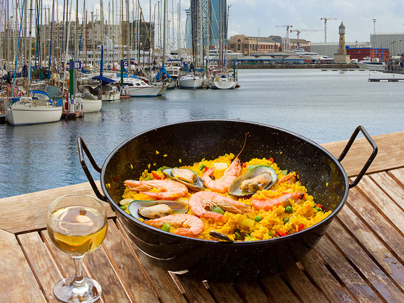 A dish of paella and a glass of wine on a table overlooking the harbor in Barcelona Spain