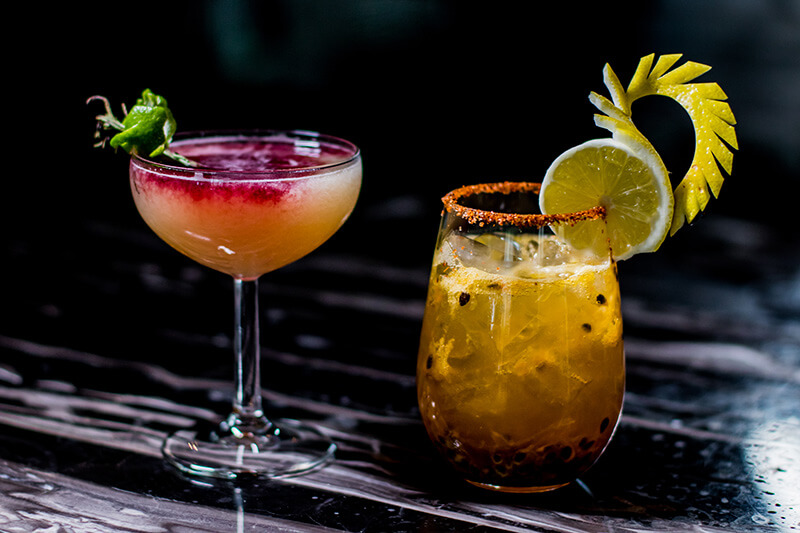 two colorful cocktails set against a black marble bar top background