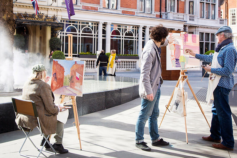 an art instructor teaching a man and a woman how to paint in front of The Connaught