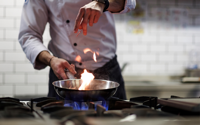 A chef in a white coat sprinkles food into a pan thats on fire