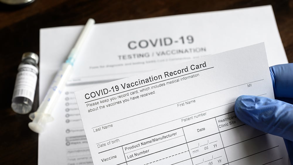 gloved hand holding COVID-19 vaccination record card with needle on table