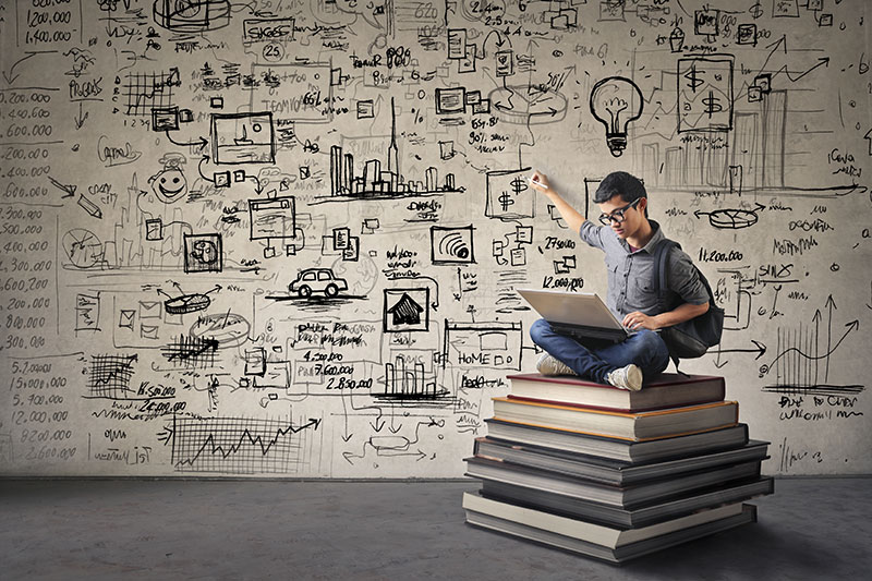 Young man with nerdy black glasses sitting on a book hill drawing his ideas creatively on the wall behind him while looking 