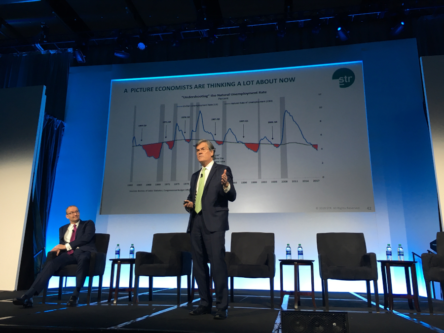 Mark Woodworth stands on stage in front of a graph showing unemployment trends