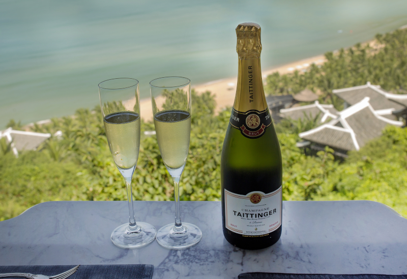 A bottle of champagne and a couple glasses sit on a table with the beach in the background