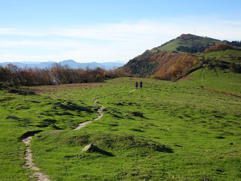Two people walking along a trail among green hills