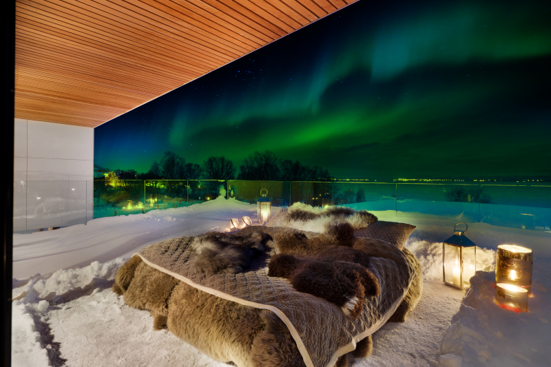 A king-sized bed sits on an outdoor balcony with the Northern Lights in the background