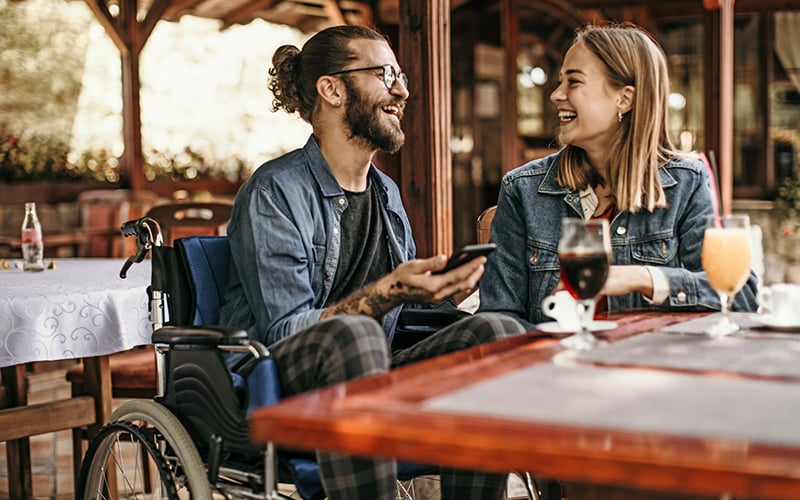 A bearded man in a wheelchair laughs with a friend at a restaurant