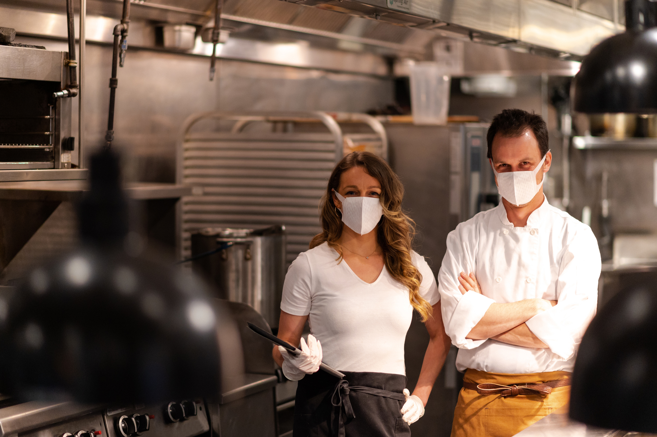 Two service industry workers wear masks inside a restaurant kitchen