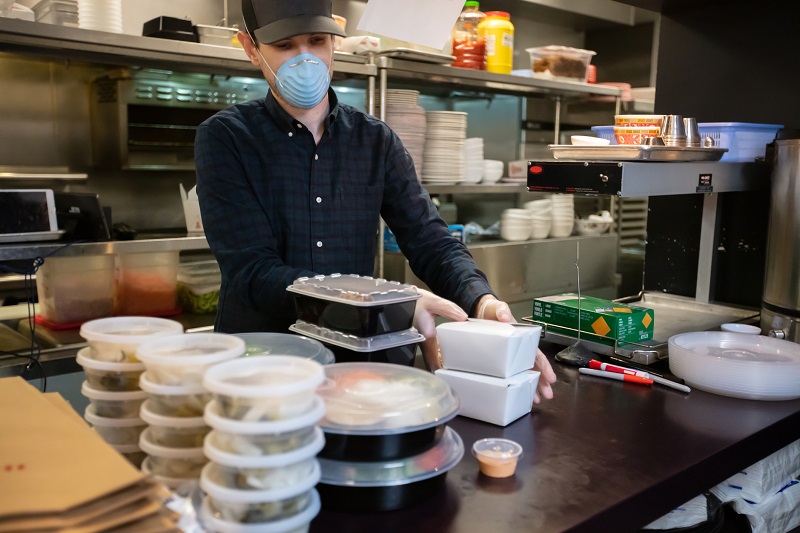 A man in a restaurant wears gloves and a mask to pack food order for delivery 