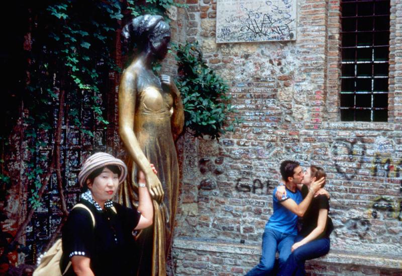 A woman standing next to a statue of Juliet, while a couple kisses behind