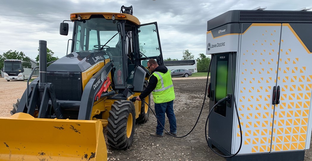 man is inserting charging cable into charging port on deere electric backhoe