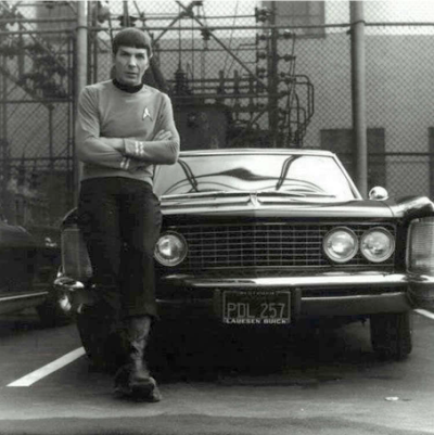 Leonard Nimoy in costume as Star Trek's Mr. Spock leaning nonchalantly against the hood of a 1960s Buick Riviera