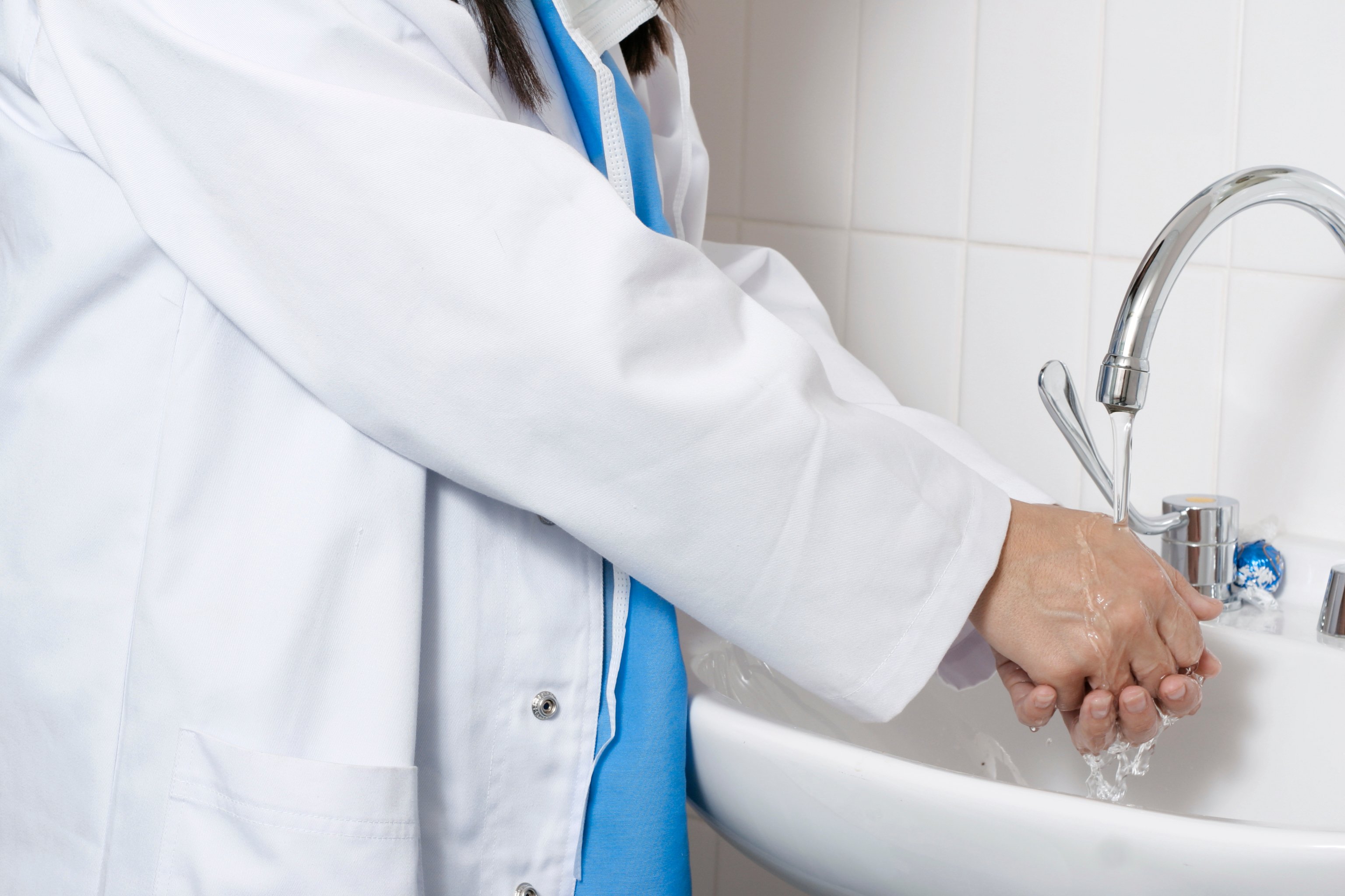 Healthcare worker in white lab coat and surgical scrubs washes his hands in sink