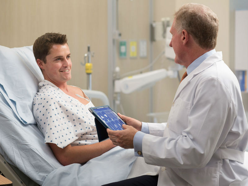 Patient wearing a VitalPatch on his chest sitting in a hospital bed while talking to a doctor looking at vital signs on a tab