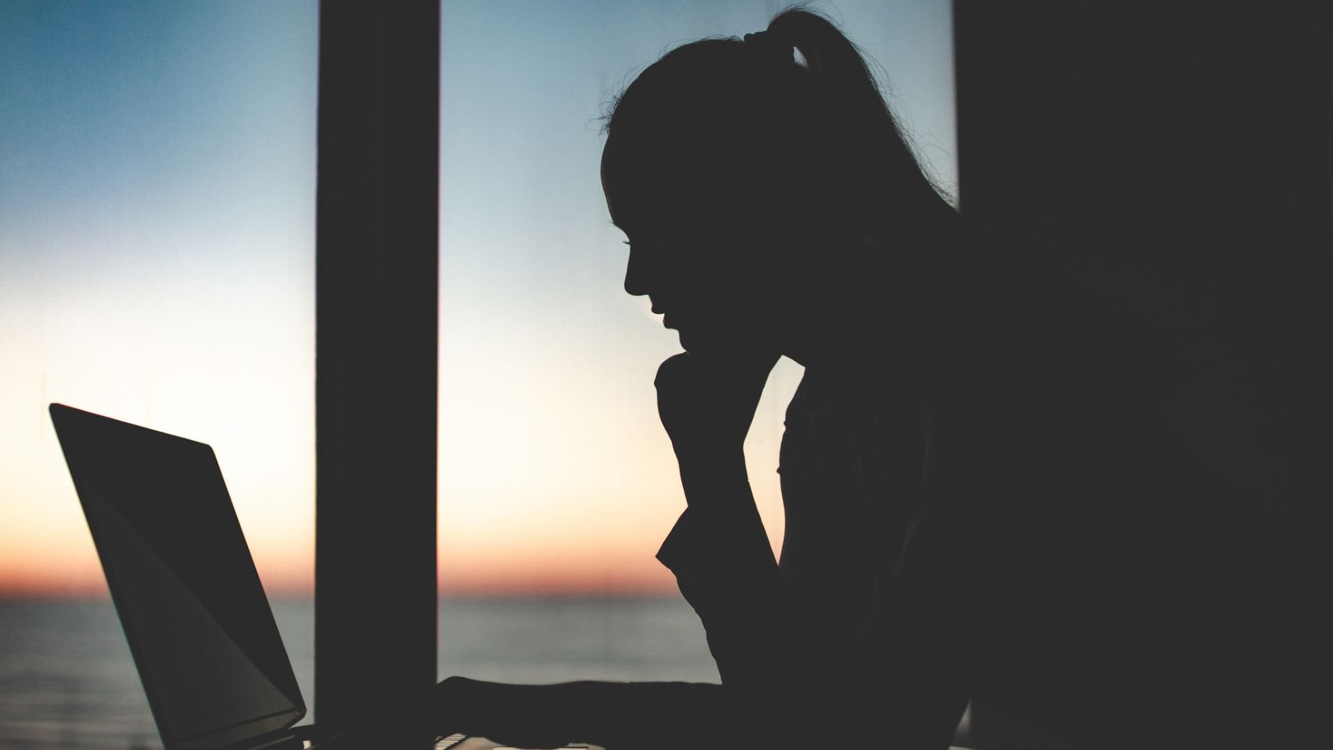 Silhouette of a business woman working late at a laptop against the window at sunset