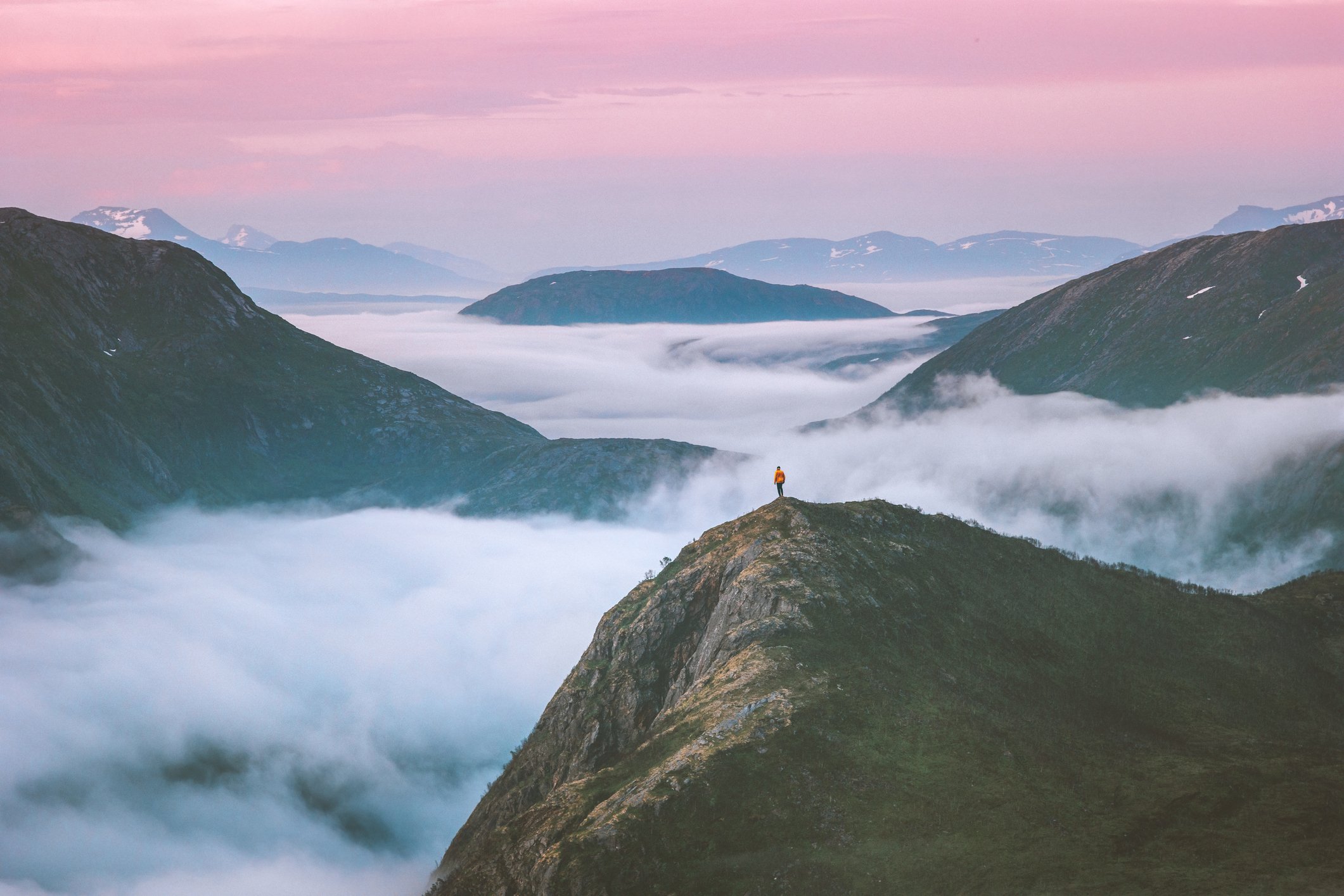 photo of a hiker on top of a mountain or hill peak look out toward the horizon over fog that cuts through other peaks