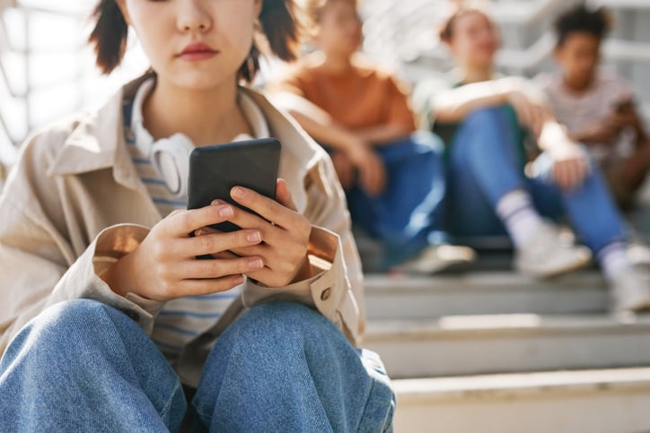 A teenage girl checks her phone while blurred teens sit in the background