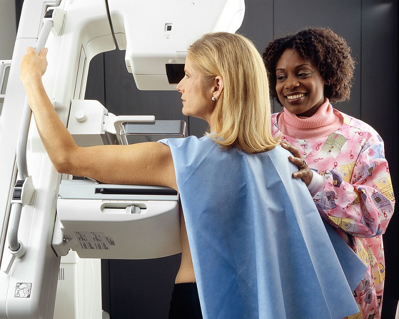 An African-American female technician positions a Caucasian woman at an imaging machine to receive a mammogram