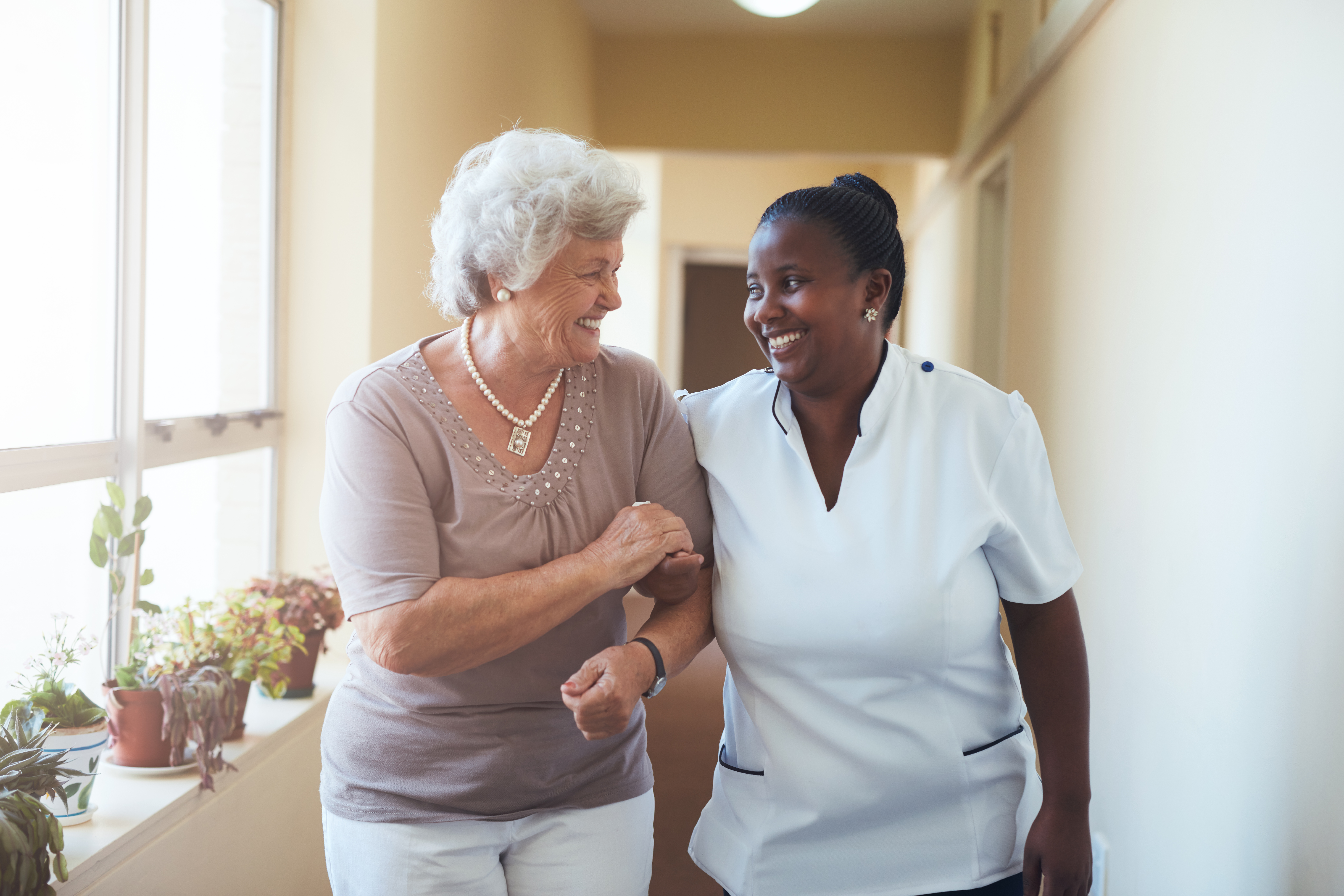 Caregiver holds arm of senior woman as they smile at one another and walk down a light-filled hallway together