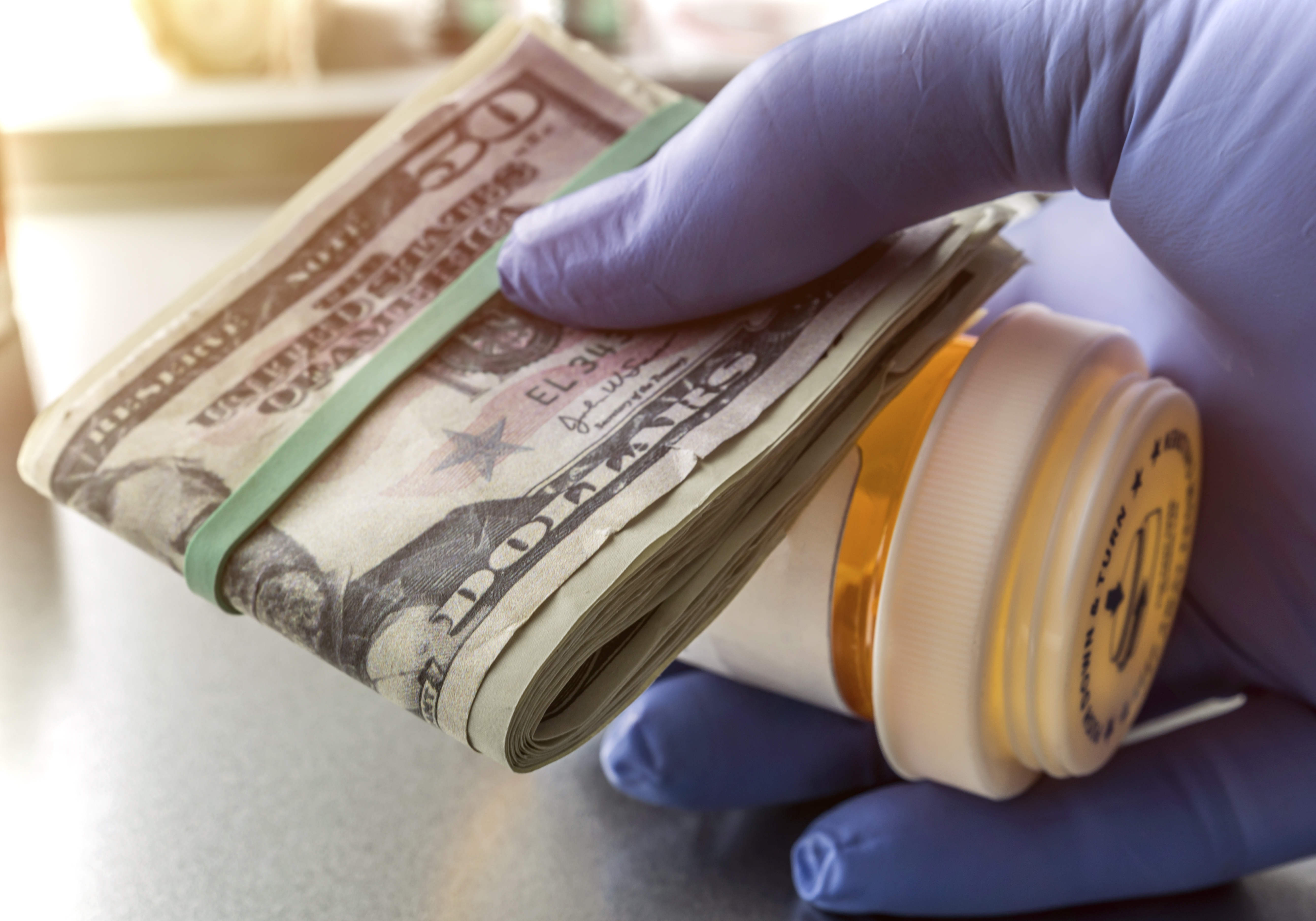 Hand wearing latex surgical glove holds prescription bottle and wad of 50-dollar bills