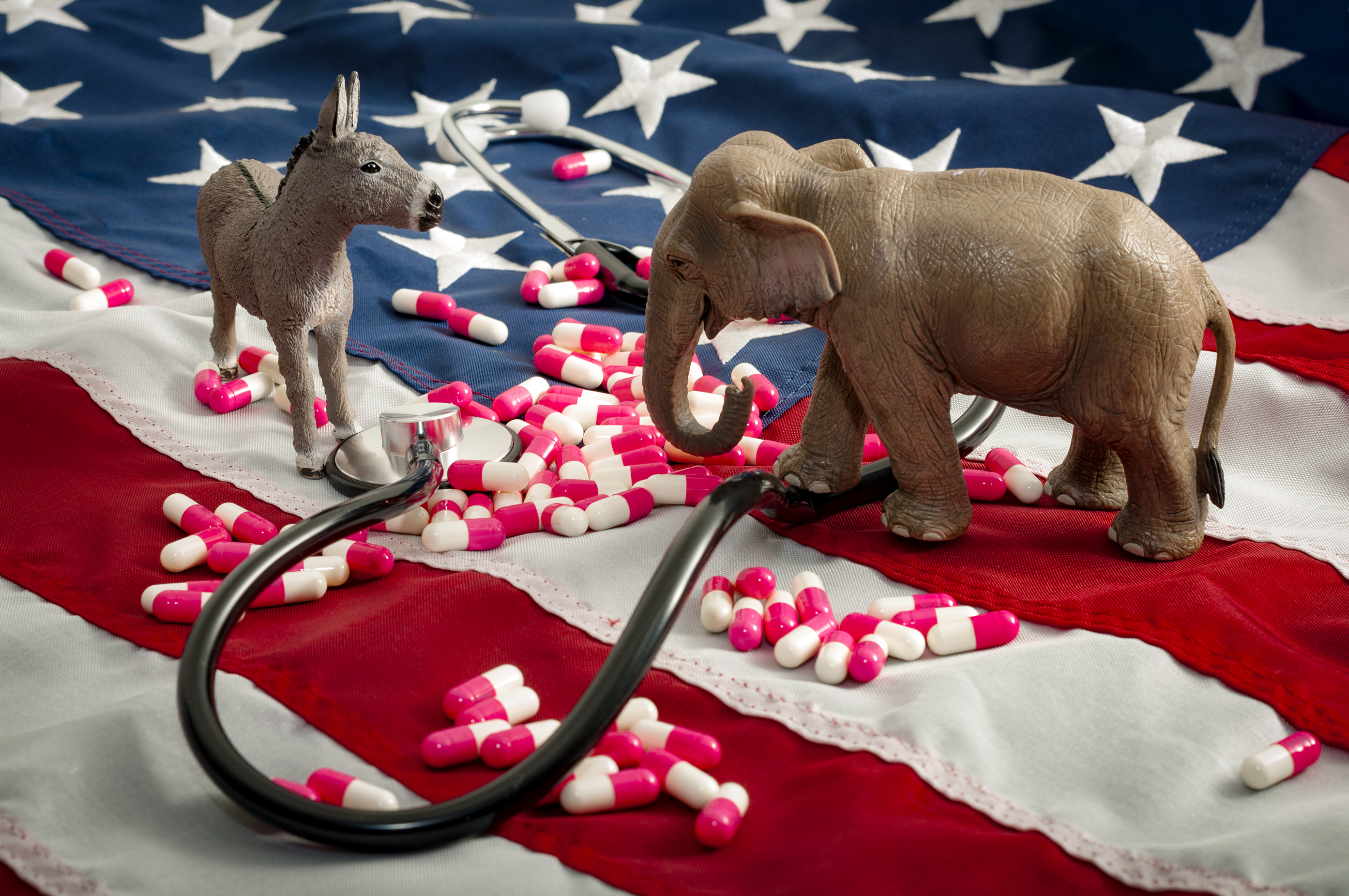 Donkey and elephant figurines face each other with stethoscope and pills with American flag in background