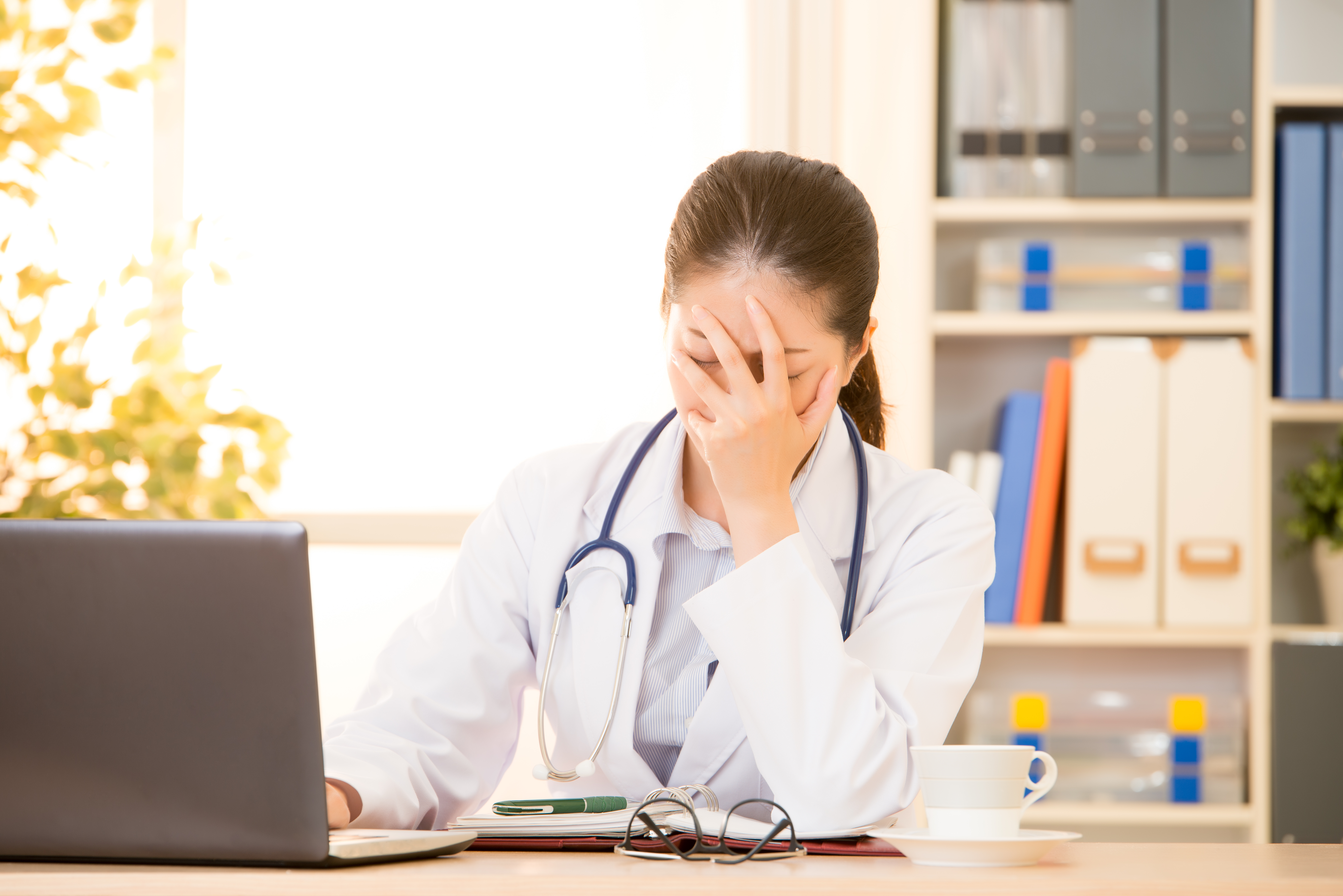 Young female doctor sitting at desk in front of computer covering face with hand in frustration