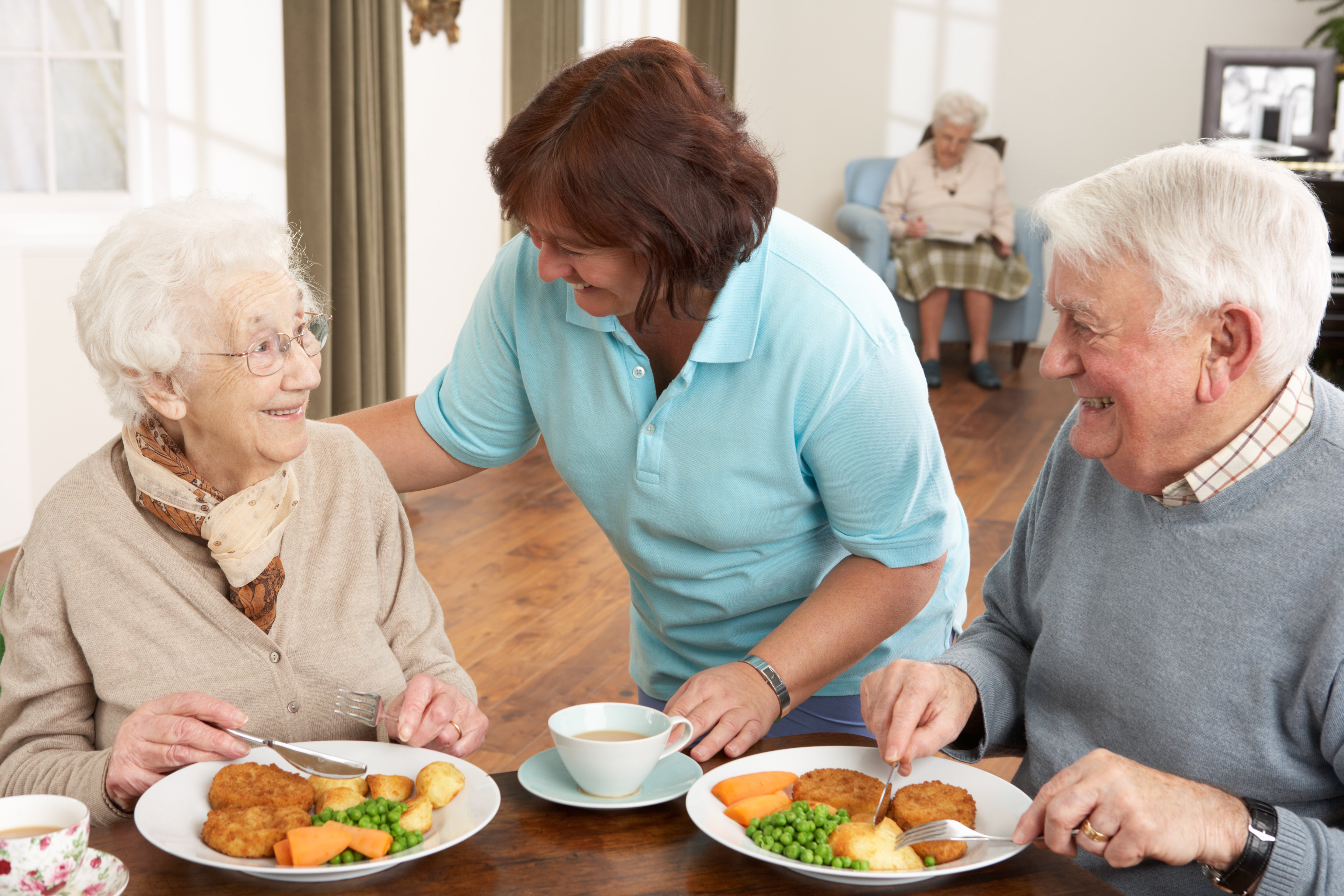 Caregiver wearing scrubs serving meal to senior man and woman at dining table