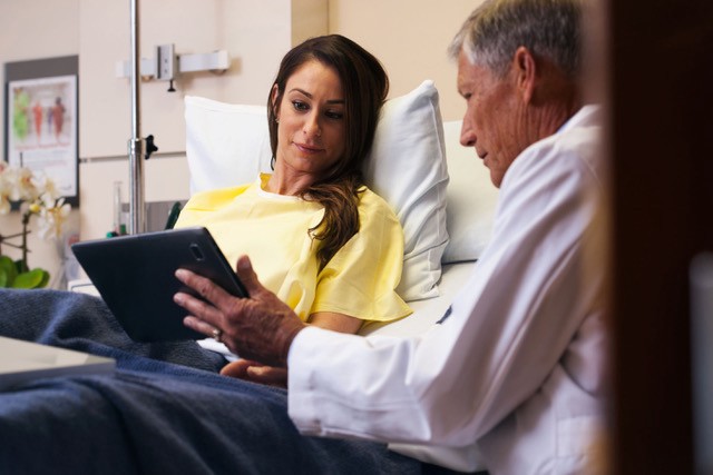 a female patient in a hospital bed speaks with a male doctor who is using a tablet to show the patient her diagnostic informa