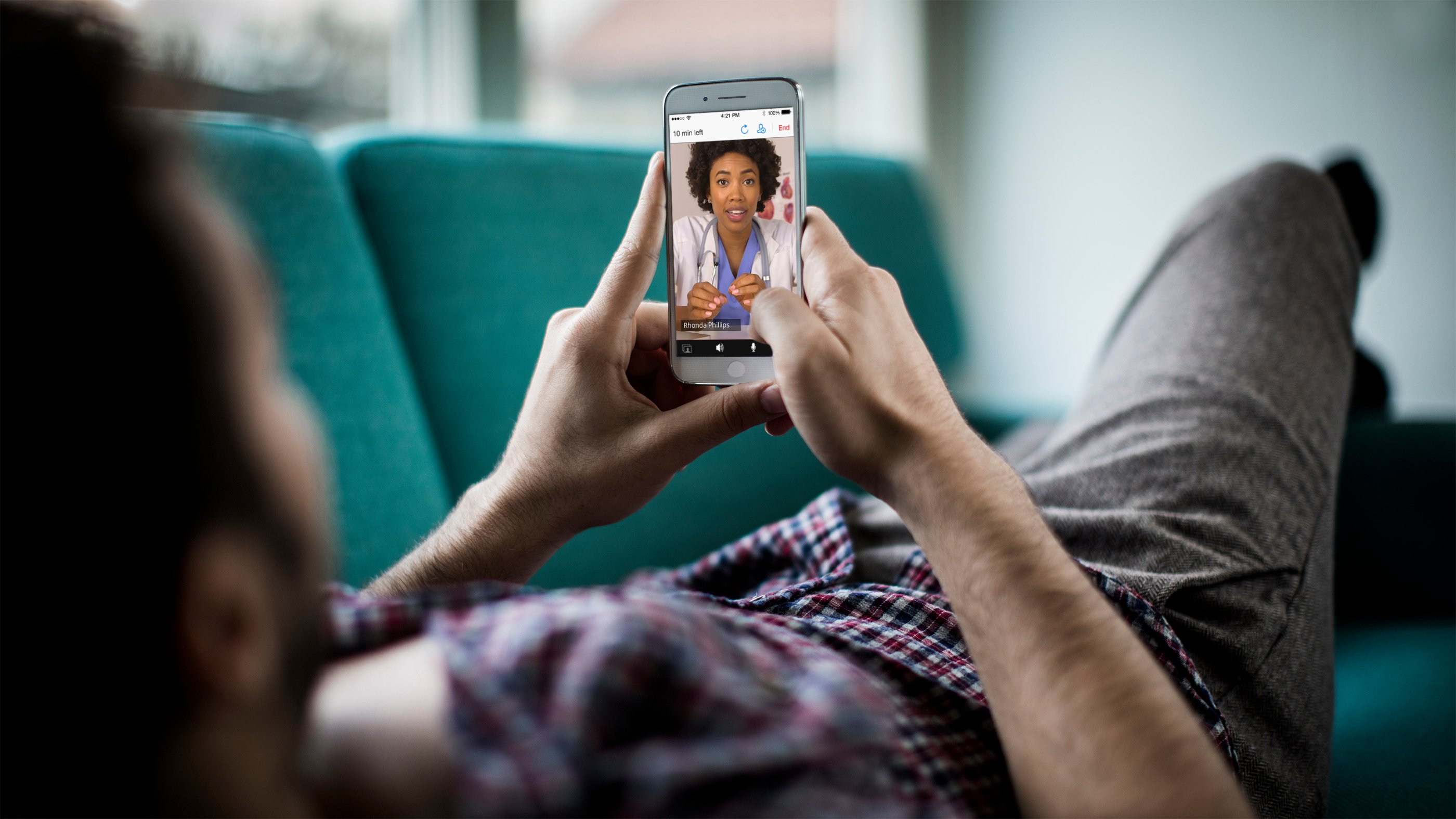 man laying on couch talks with a female doctor using a telehealth app on his smartphone