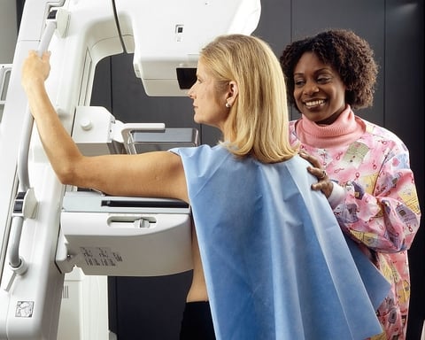 An African-American female technician positions a Caucasian woman at an imaging machine to receive a mammogram.