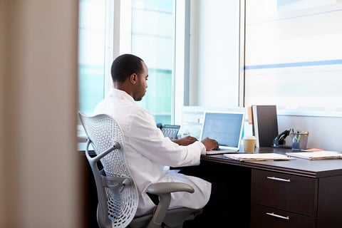 A doctor sitting at his desk working on a laptop computer.