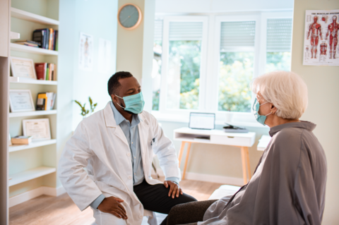 Primary care doctor A male primary care doctor and his patient sit across from each other talking. Both are wearing masks