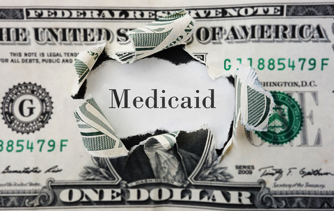 Dollar bill with a hole in Washington's face on it and the word "Medicaid" in its place