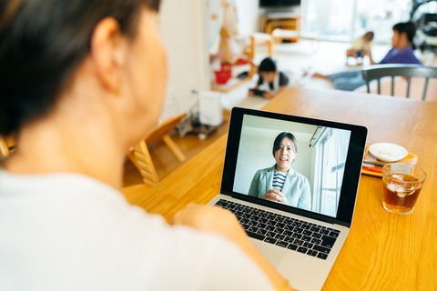 A mother is talking with a mental health professional via web conference at home while her children are playing in the background.