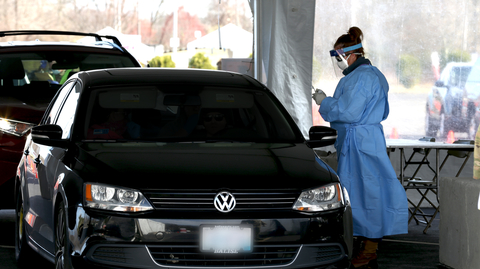 healthcare worker in protective gear prepares to test a driver in a car at a drive-through COVID testing site