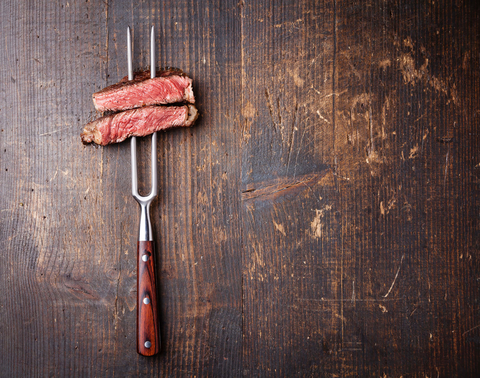 Slices of beef on meat fork against dark wood background Slices of beef steak on meat fork on dark wooden background