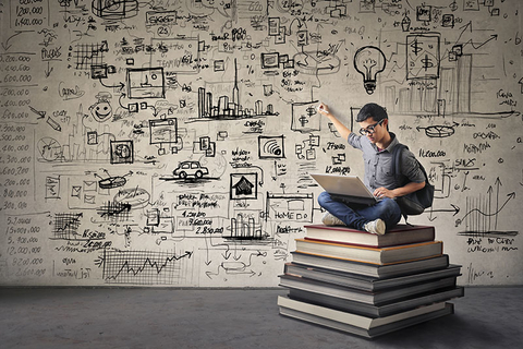 Business Inspiration Getty Young man with nerdy black glasses sitting on a book hill, drawing his ideas creatively on the wall behind him while looking at his computer.