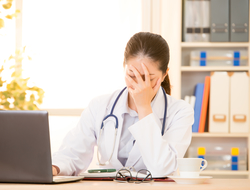 Frustrated doctor Young female doctor sitting at desk in front of computer covering face with hand in frustration